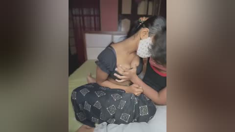 Media: Video of a woman with medium-dark skin and medium breasts being breastfed by an older man with light skin and gray hair. They are in a dimly lit bedroom with a bed, shelves, and a red wall in the background.