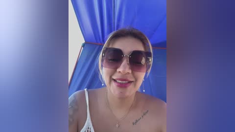 Media: Video of a smiling woman with light skin, light brown hair, and large sunglasses, wearing a white spaghetti strap top. She has a tattoo on her left shoulder.