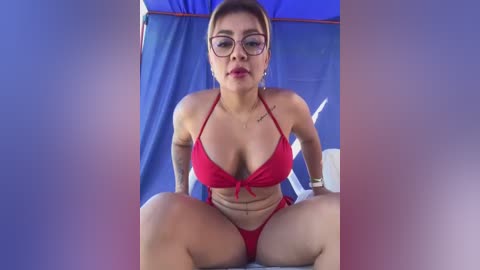 Media: Video of a Latina woman with light skin, medium build, and short brown hair, wearing glasses and a red bikini, seated indoors with blue curtains in the background.