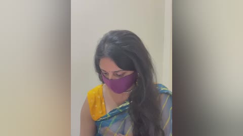 Media: A video of a South Asian woman with long black hair, wearing a yellow blouse, blue and yellow saree, and a purple face mask, standing indoors against a plain white wall.