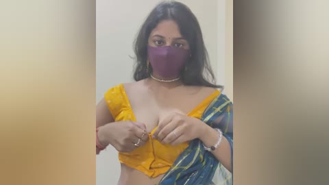 Media: Video of a South Asian woman with long dark hair, wearing a yellow blouse and blue sari, purple mask, and a pearl necklace.