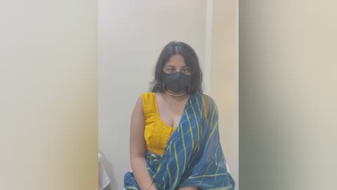 Media: Video of a young woman with medium skin tone and black hair, wearing a yellow blouse, blue sari with green stripes, and black face mask, sitting in a white-walled, narrow hallway.