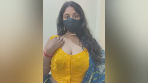 Media: Video of a South Asian woman with long black hair, wearing a yellow polka-dotted blouse, blue sari, black face mask, and a red bracelet, against a blurred beige wall.