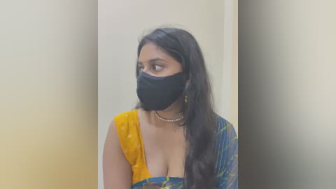 Media: Video of a South Asian woman with long black hair, wearing a yellow and blue saree, black face mask, and a pearl necklace, standing against a beige wall, looking to the right.