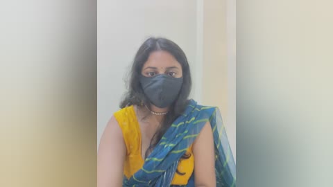 Media: A video of a woman with medium brown skin, wearing a yellow blouse, blue sari with green stripes, and a black mask, standing against a plain beige wall.