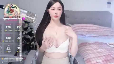 Media: A video of a young East Asian woman with long black hair, wearing a white bra and beige pants, sitting on a grey couch with a pink blanket. The background shows a gaming setup with a computer and a plush toy.