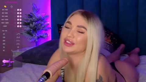 Media: Video of a blonde woman with closed eyes, lying on a bed, being pleasured by a man's hand. Background features a teal curtain and a purple-lit Christmas tree.