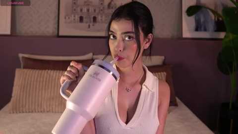 Video of a young woman with dark hair in a white sleeveless top, blowing into a white hair dryer. She's in a bedroom with brown walls and framed art.