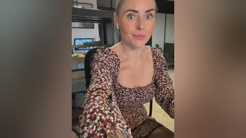 Media: Video of a fair-skinned woman with light brown hair, wearing a floral-patterned dress, seated in an office with a desk and computer monitor in the background.