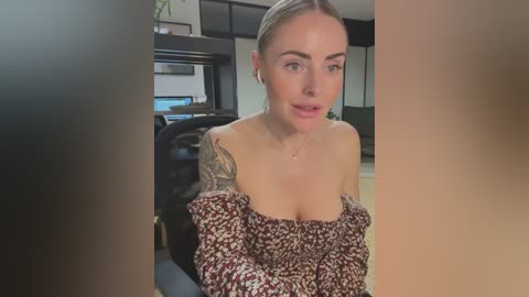 Media: Video of a blonde woman with fair skin and a tattoo on her right shoulder, wearing a off-the-shoulder floral dress, seated indoors, blurred background.