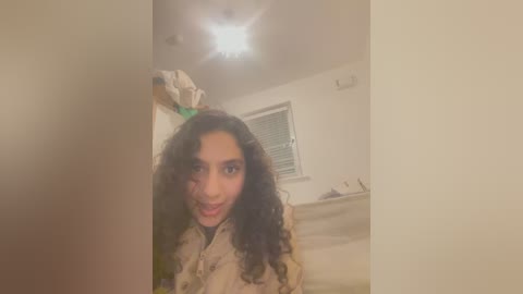 Media: Video of a young woman with curly black hair, wearing a beige jacket, playfully holding a green object above her head, in a dimly lit room with a window and a ceiling light.