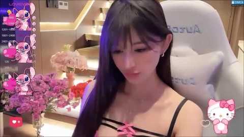 Media: Video of an Asian woman with long black hair, wearing a black lingerie top with pink bows, sitting in a plush chair in a modern room.