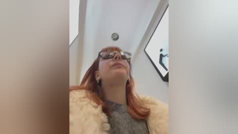 Video of a young woman with fair skin, long red hair, and glasses, wearing a grey sweater and beige fur coat, captured from a low angle inside a white-walled elevator.