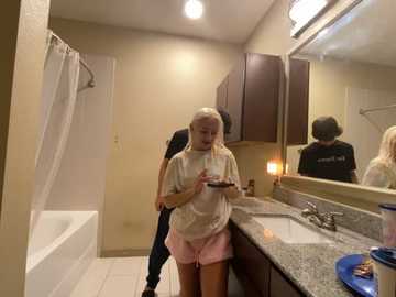 Media: Video of an elderly woman with long blonde hair in a beige t-shirt and pink shorts, looking at a bowl of food, in a beige-tiled bathroom with a bathtub, sink, and mirror.