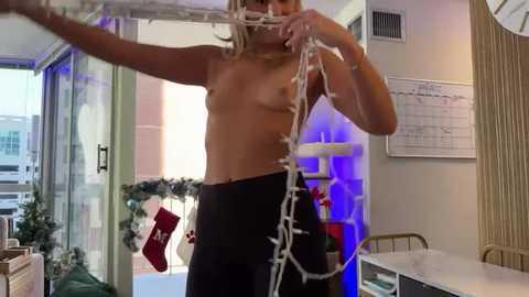 Media: Video of a topless woman with blonde hair, wearing black leggings, stringing white Christmas lights in a modern, brightly-lit room with windows, Christmas decorations, and a calendar on the wall.