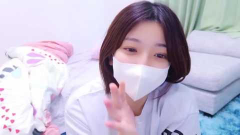 Media: Video of a young Asian woman with straight, dark hair and light skin, wearing a white surgical mask, gesturing with her right hand. She's indoors, next to a bed with a colorful, patterned blanket and a green pillow.