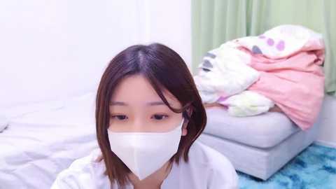 Media: Video of a young Asian woman with straight, shoulder-length brown hair, wearing a white face mask, sitting on a bed with messy white and pink sheets in a simple, brightly lit room.