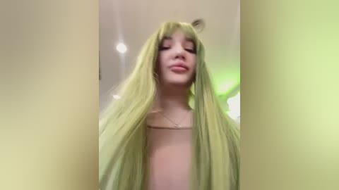 Media: A video of a fair-skinned woman with long, straight, light green hair, wearing a light green top. She has a soft, serene expression with closed eyes. The background is a blurred, indoor setting with soft lighting.