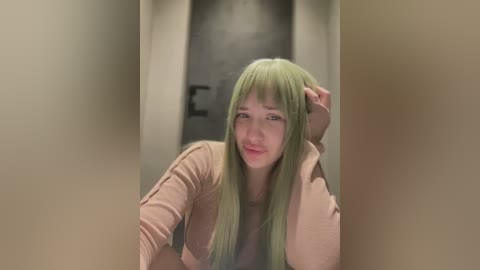 Media: Video of a young woman with long, straight, mint-green hair, wearing a beige top, leaning against a beige wall in a dimly lit corridor. She has a contemplative expression, her left hand resting on her head.