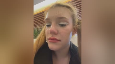 Media: Video of a young, pale-skinned woman with long blonde hair in a ponytail, wearing black clothing, and heavy white eyeliner, looking tired and contemplative. Background includes horizontal blinds and a green plant.