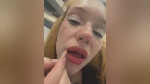 Media: Video of a fair-skinned, red-haired young woman with a nose ring, applying lipstick with a red-tipped finger. The background shows vertical black and white stripes.