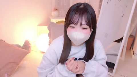 Video of an Asian woman with long black hair and a white mask, sitting on the floor in a softly lit room with a warm, cozy ambiance.