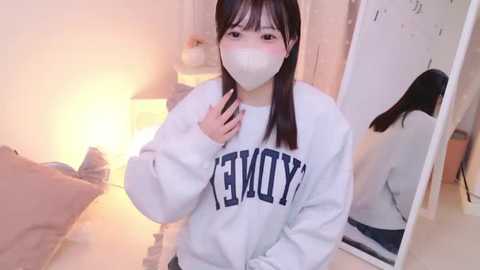 Media: Video of a young East Asian woman with long black hair, wearing a white sweatshirt with \"YAMA\" in blue letters, a face mask, and a mirror reflection in a softly lit, cozy bedroom.