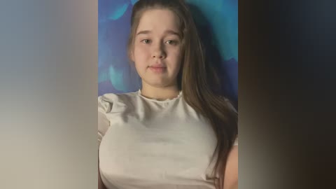 Media: A video of a young Caucasian girl with long brown hair and light skin, wearing a white blouse, against a blue background.