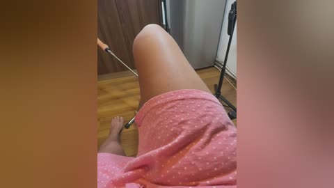 Media: A video captures a person reclining on a wooden floor, wearing pink polka-dot shorts and a pink top. The background includes a wooden door and a silver metal stand. The image is slightly blurred, with a warm, brownish tint.