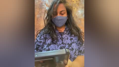 Media: Video of a middle-aged man with medium-brown skin and long, wavy black hair, wearing a blue surgical mask and a dark blue floral shirt. He is holding a black tablet computer in front of a beige tiled wall.