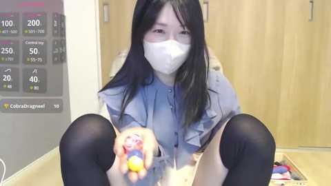 Media: A video of a young Asian woman with long black hair, wearing a white surgical mask, blue denim shirt, black knee-high socks, and sitting on the floor. She holds a colorful plastic toy in front of a wooden wardrobe and a digital display.