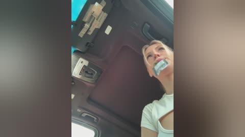 Media: Video of a woman with blonde hair, gagged with a blue and white cloth, seated in the backseat of a car, wearing a white T-shirt, surrounded by clutter.