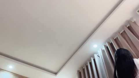 Media: Video of a ceiling with recessed lighting, beige curtains, and a person with long black hair standing in the foreground, possibly looking out a window.