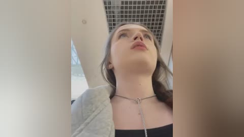 Media: Video of a young Caucasian woman with fair skin, brown hair, and a black top, wearing a silver chain necklace. She gazes upward. Background features a grid of square tiles and a white ceiling.