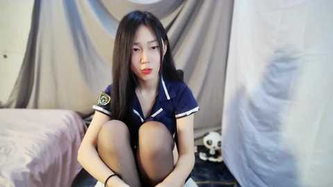 Media: Video of a young East Asian woman with long black hair, wearing a navy blue sailor outfit, sitting on a bed with a white sheet and a stuffed panda toy.