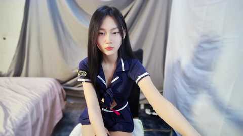 Media: Video of an Asian woman in a navy blue, short-sleeved sailor's outfit with a red tie, sitting on a bed with grey curtains in the background.