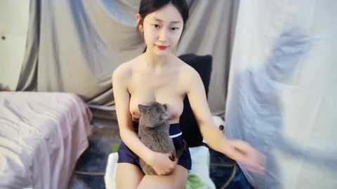 Media: Video of a topless Asian woman with medium-sized breasts, dark hair, and fair skin, seated on a bed holding a gray cat. She wears a blue skirt. Background features a draped, neutral-colored fabric.