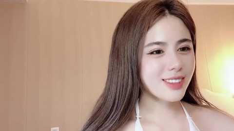 Media: Video of a young Asian woman with long, straight brown hair, fair skin, and a warm smile. She wears a white halter top, set against a beige wall.