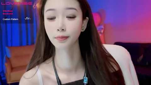 Media: A video of an East Asian woman with long, straight black hair, closed eyes, and a serene expression, wearing a black top and necklace, in a softly lit room with blue and red lighting.