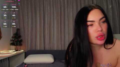 Media: Video of a topless woman with long black hair, wearing a necklace, in a dimly lit room with grey curtains, a small Christmas tree, and a white pillow.