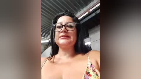 Media: Video of a fair-skinned woman with dark hair, wearing glasses and a colorful floral top, standing in a warehouse with metal beams and exposed pipes.