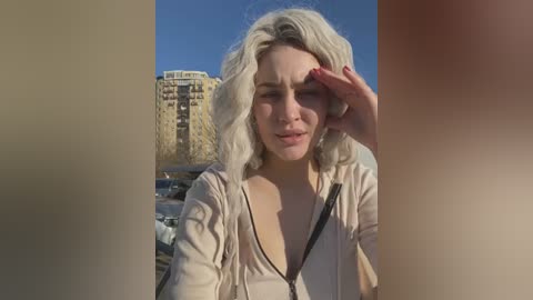 Media: A video of a blonde woman with shoulder-length hair, wearing a beige top, shielding her eyes from the sun. Background features a beige high-rise building under a clear blue sky.