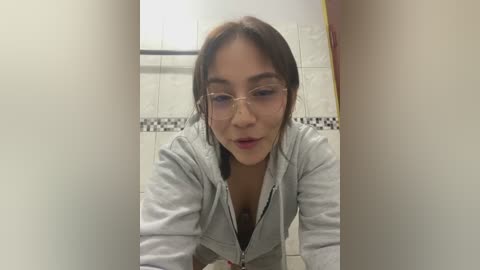 Media: Video of a young woman with light skin and brown hair, wearing glasses, leaning forward in a restroom with white tiled walls and a black and white patterned border.