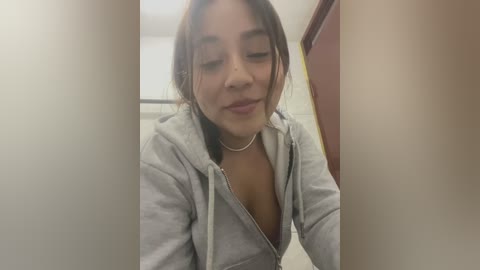 Media: Video of a young woman with medium brown skin, medium-length brown hair, and closed eyes. She wears a light grey hoodie with a zipper open, revealing a hint of cleavage. The background features a beige wall and a door.