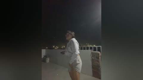 Media: A video of a woman in a white robe and short shorts, standing on a rooftop at night, illuminated by a bright light.