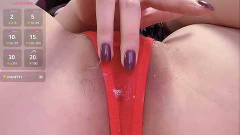 A close-up video of a woman's hand pulling aside a red thong, revealing her pubic area with visible pubic hair. The background is blurred, focusing on the intimate act.