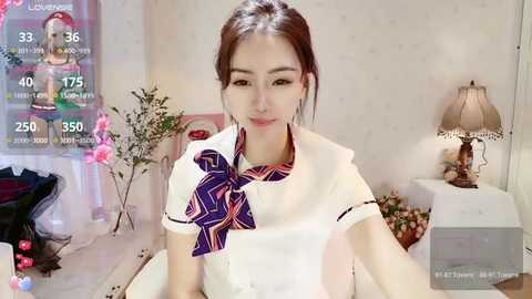 Media: A video of an East Asian woman with fair skin, straight brown hair, and a white blouse with a purple bow. She's in a cozy, well-lit room with a white lamp and pink flowers.