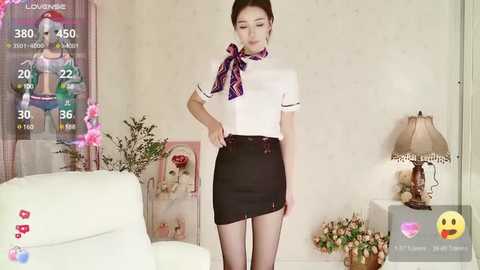 Media: Video of a slender East Asian woman in a white blouse with a purple bow and black mini skirt, standing in a cozy, beige-themed living room with a white couch, a lamp, and a poster.