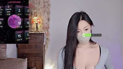 Media: Video of a young Asian woman with long black hair, wearing a surgical mask, in a dimly lit bedroom with a wooden dresser, lamp, and a wall calendar displaying a lunar eclipse.