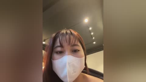 Media: Video of a young Asian woman with long brown hair and bangs, wearing a white face mask, taken from a low angle, indoors with dim lighting and ceiling lights.